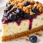 Blueberry crumble cheesecake slice with creamy cheesecake, fresh blueberries, and golden crumble topping on a white plate.