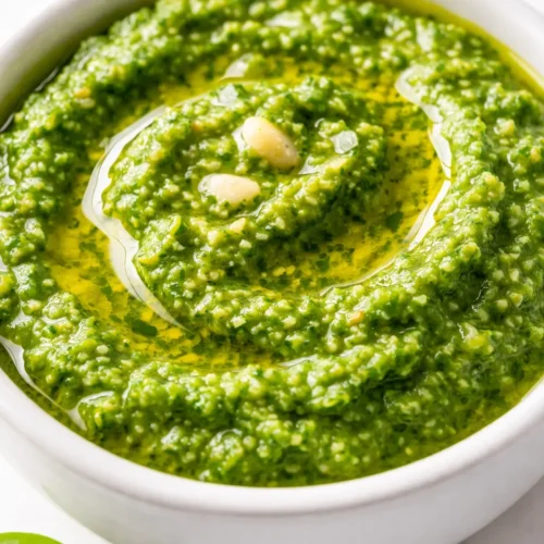 Vibrant green fresh basil pesto spooned over pasta, with glossy texture and visible basil leaves and pine nuts.