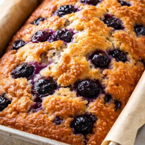 Moist blueberry quick bread loaf with golden crust and plump blueberries, sliced on a wooden board.