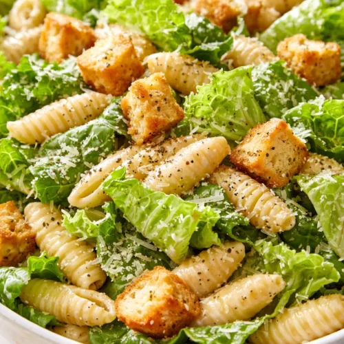 Creamy chicken Caesar pasta salad with crunchy croutons, parmesan shavings, and crisp romaine lettuce in a wooden bowl.