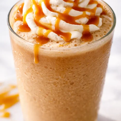 Tall glass of DIY Caramel Crunch Frappe topped with whipped cream, caramel drizzle, and crunchy bits, creamy brown tones.