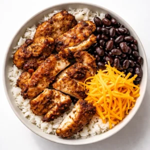 Assembling a high protein chicken burrito bowl with grilled chicken, black beans, cilantro lime rice, corn, avocado, and a drizzle of crema.