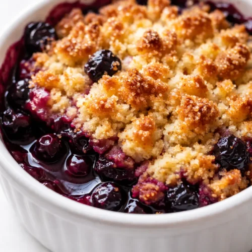 Golden strawberry shortcake crumble topping on a deep purple huckleberry pie, with a sliced piece revealing juicy filling.