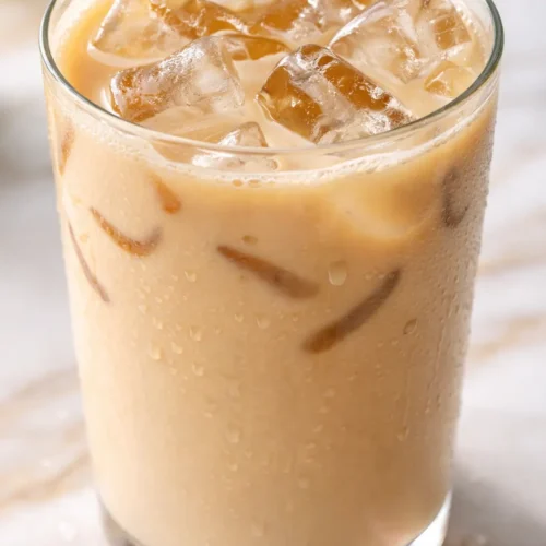 Tall glass of iced vanilla chai latte with creamy milk, ice cubes, and a sprinkle of cinnamon on top.