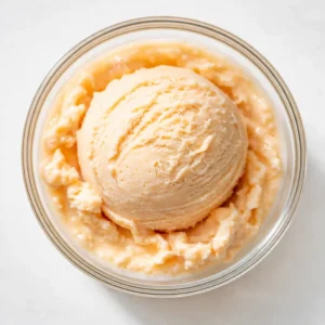 Creamy peach ice cream base being stirred with chunks of ripe peaches, showing smooth texture and vibrant orange color.