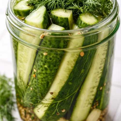 Crisp, bright green dill pickles in a glass jar with garlic cloves, fresh dill sprigs, and a clear brine.