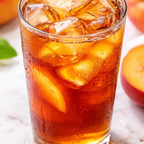 Tall glass of refreshing peach iced tea with golden amber color, peach slices, and ice cubes, garnished with mint leaves.