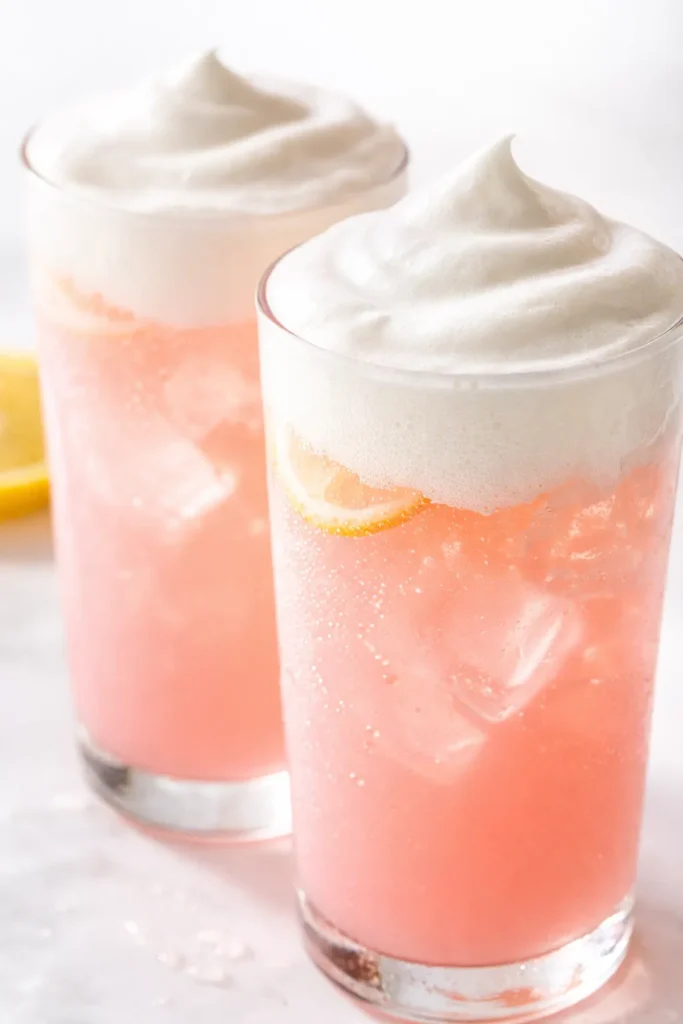 Tall glass of whipped pink lemonade with a thick white foam layer on top, garnished with a lemon slice.