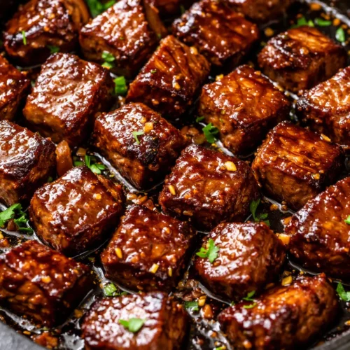 Glossy honey garlic steak bites with caramelized edges in a shiny sauce, topped with sesame seeds and green onions.